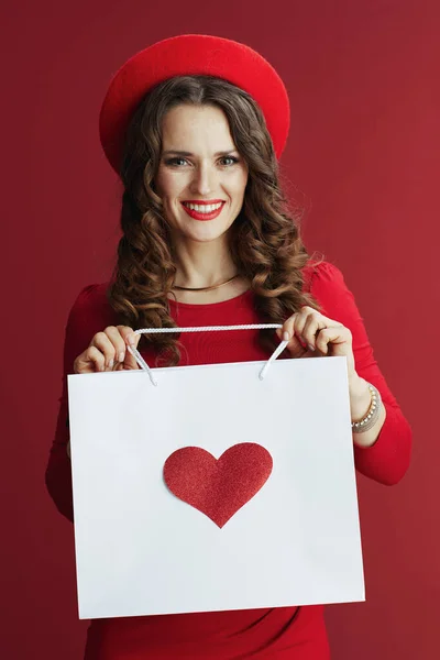 ハッピーバレンタイン スタイリッシュな40歳の女性を笑顔で赤いドレスとベレー帽で隔離された赤の背景にショッピングバッグ付きハート — ストック写真