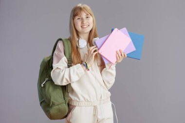 Gri arka planda izole edilmiş sırt çantası, çalışma kitabı ve kulaklığı olan bej eşofmanlı mutlu modern kız portresi.