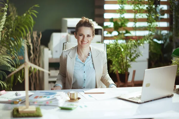 Portrait of happy modern accountant woman in a light business suit in modern green office with laptop.