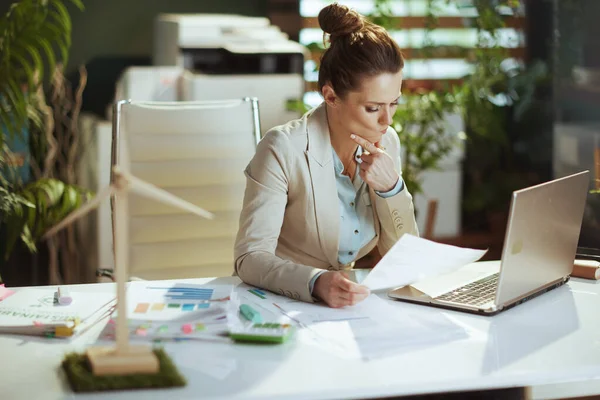 modern middle aged accountant woman in a light business suit in modern green office with documents and laptop.