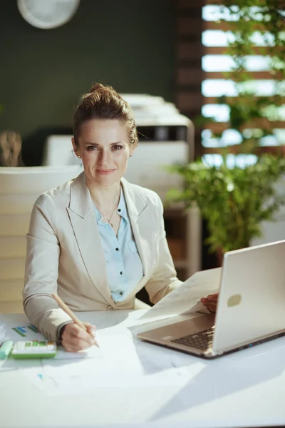 Portrait of modern bookkeeper woman in a light business suit in modern green office with documents and laptop.