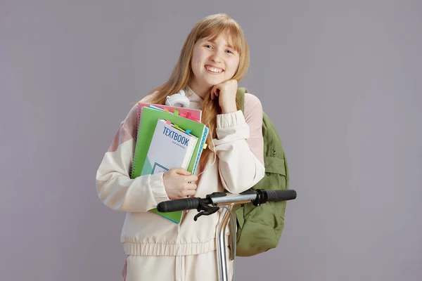 幸せなトレンディな女の子でベージュトラックスーツでバックパック ワークブック スクーターや教科書とグレーの背景に隔離 — ストック写真