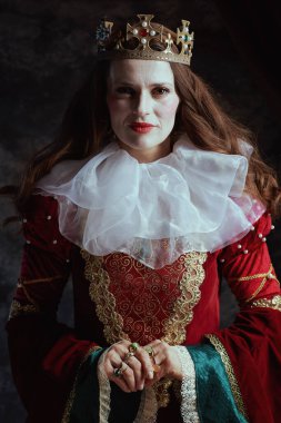 Kırmızı elbiseli, beyaz yakalı ve tacı koyu gri arka planlı ortaçağ kraliçesi..
