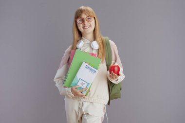 Elma, sırt çantası, çalışma kitabı, kulaklık, ders kitabı ve gözlüklü bej eşofmanlı mutlu bir genç kız..