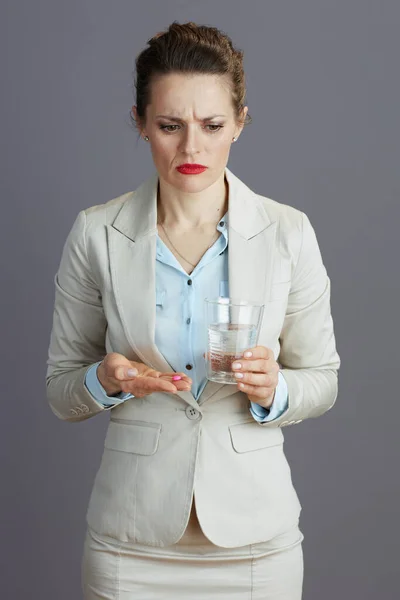 穿着浅色西服的疲惫而优雅的女工 带着一杯水和药丸 与灰色隔离 — 图库照片