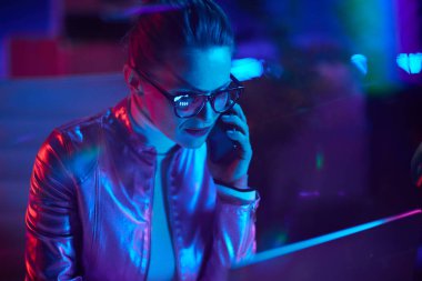 Neon metaevren fütüristik konsepti. Düşünceli, 40 yaşında, gözlüklü, laptoplu, modern ofisteki akıllı telefondan konuşan bir kadın..