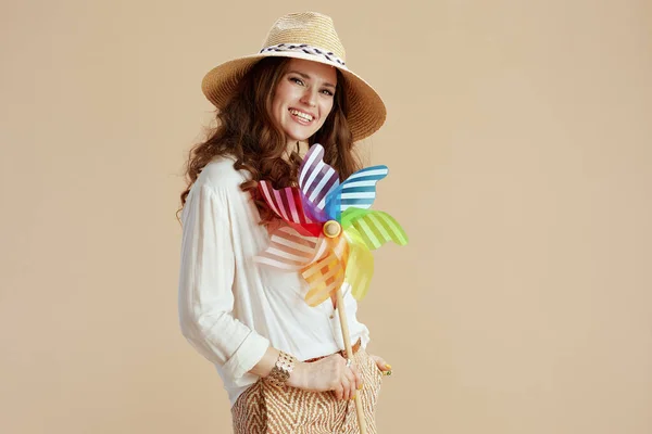 ビーチ休暇 笑顔現代的な中年主婦でホワイトブラウスとショートパンツにベージュ背景とともに風車のおもちゃと夏の帽子 — ストック写真