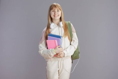 Sırt çantası, çalışma kitabı ve kulaklığı gri renge karşı olan bej eşofmanlı, güler yüzlü genç kadının portresi.