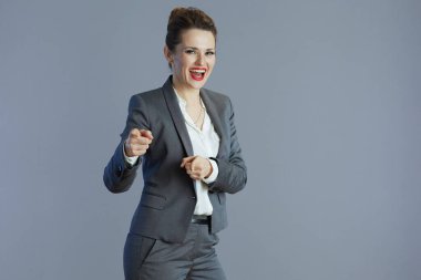 Gri takımlı modern orta yaşlı kadın işçi gri üzerine izole edilmiş kamerayı işaret ediyor..