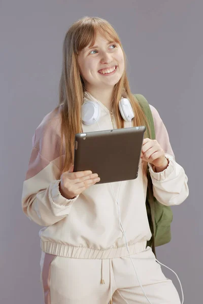 幸せなトレンディーなティーン女の子でベージュトラックスーツでバックパック ヘッドフォンとタブレットPc対グレー — ストック写真