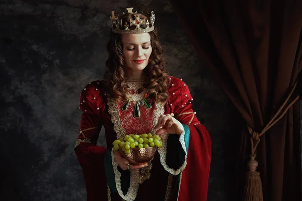 快乐的中世纪女王 身穿红色衣服 有一盘葡萄 头戴深灰色背景的皇冠 — 图库照片