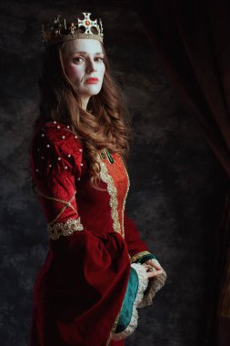 Kırmızı elbiseli, beyaz makyajlı ve tacı koyu gri arka planda olan ortaçağ kraliçesi..