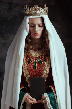 Kırmızı elbiseli, kitap, peçe ve tacı koyu gri arka planda olan ortaçağ kraliçesi.
