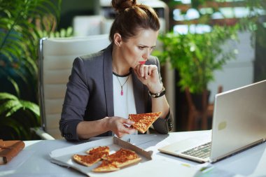 Sürdürülebilir iş yeri. Modern yeşil ofisteki gri takım elbiseli, pizza ve dizüstü bilgisayarlı modern muhasebeci kadınla ilgiliydi..