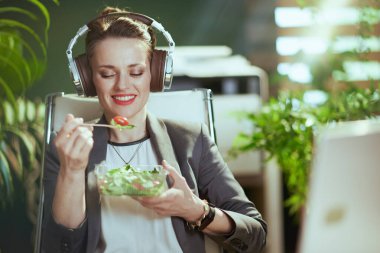 Sürdürülebilir iş yeri. Modern yeşil ofisteki gri takım elbiseli, modern 40 yaşındaki gülümseyen muhasebeci kadın. Salata yiyor ve kulaklıkla müzik dinliyor..