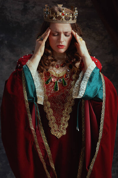 stressed medieval queen in red dress with crown having headache on dark gray background.