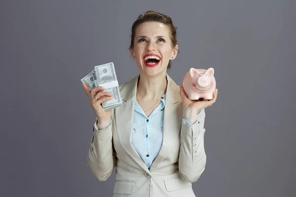 スタイリッシュな女性労働者を笑顔でピンクの銀行とドルのお金パックで軽いビジネススーツグレーの背景に隔離された — ストック写真