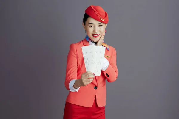 smiling stylish asian female air hostess in red skirt, jacket and hat uniform with flight tickets against grey background.