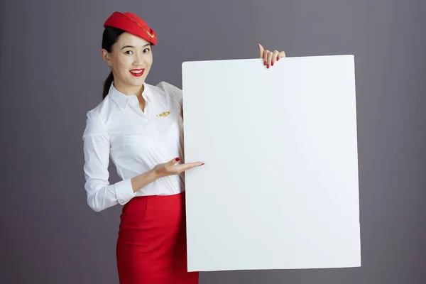 快乐的现代亚洲女乘务员 身穿红色短裙 头戴帽子制服 空白广告牌 灰色相间 — 图库照片
