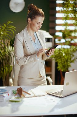 Modern yeşil ofisteki hafif takım elbiseli, dizüstü bilgisayarı ve akıllı telefonu olan mutlu, küçük işletme sahibi kadın..