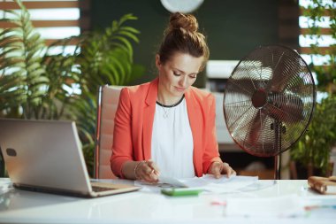 Sürdürülebilir iş yeri. Modern 40 yaşındaki muhasebeci kadın, kırmızı ceketli, elektrikli yelpaze ve dizüstü bilgisayarlı, modern yeşil ofiste çalışıyor..