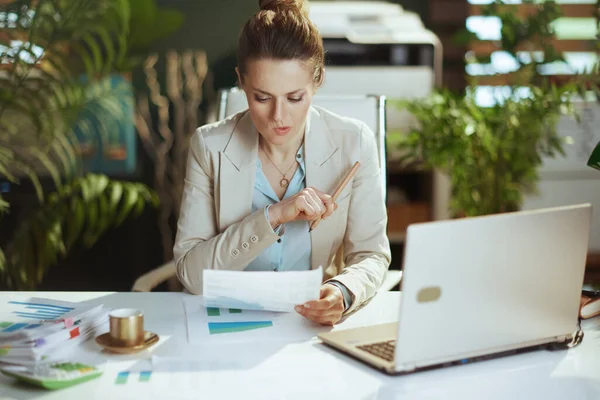 現代の40歳の女性従業員書類やノートパソコン付きのモダンなグリーンオフィスで軽いビジネススーツを着て — ストック写真