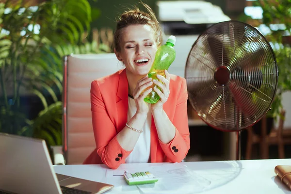 Sustainable workplace. smiling modern middle aged accountant woman in modern green office in red jacket with bottle of water, electric fan and laptop.