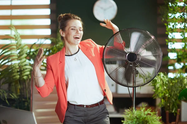 持続可能な職場 幸せな現代40歳の女性従業員で現代緑のオフィスで赤のジャケットで電気ファン — ストック写真