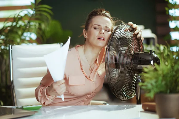 stock image Sustainable workplace. modern middle aged business woman at work with documents and electric fan suffering from summer heat.