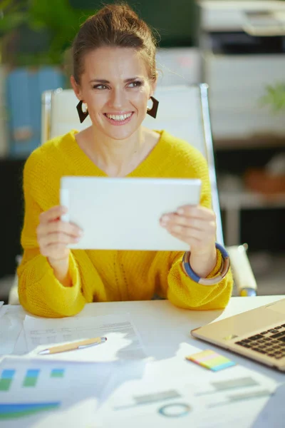 幸せな中年中小企業所有者女性で黄色セーターでタブレットPcで現代の緑のオフィス — ストック写真