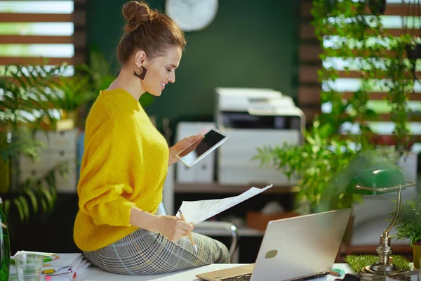幸せな中年中小企業所有者女性で黄色のセーターでラップトップを使用してタブレットPcと現代の緑のオフィスで文書を扱う — ストック写真