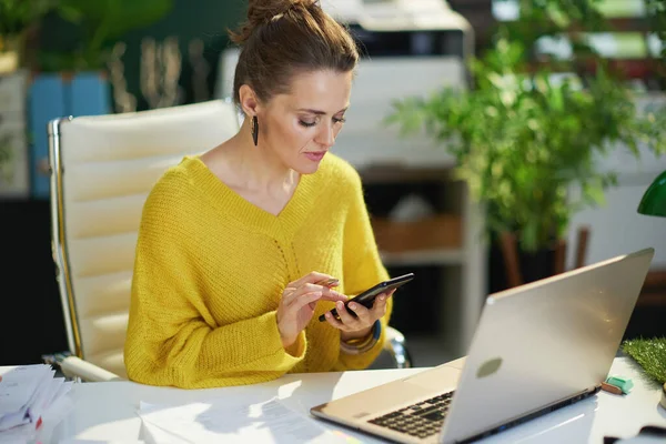 在现代绿色办公室里 穿着黄色毛衣 手持智能手机和笔记本电脑的现代小业主妇女 — 图库照片