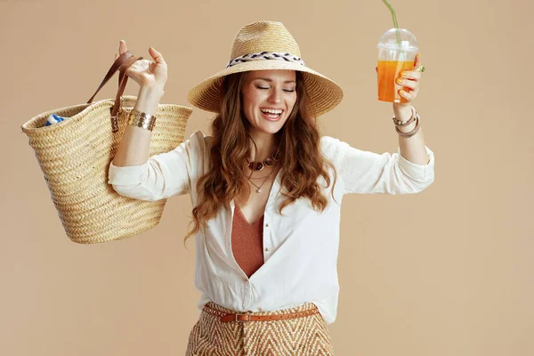 ビーチ休暇 幸せなスタイリッシュな中年主婦でホワイトブラウスとショートパンツ隔離上のベージュの背景にストローバッグ ニンジンスムージーと夏の帽子 — ストック写真