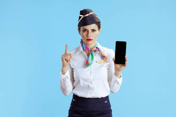 蓝色背景的现代女性空姐 穿着制服 显示智能手机空白屏幕 有一个凸起的手指吸引注意 — 图库照片