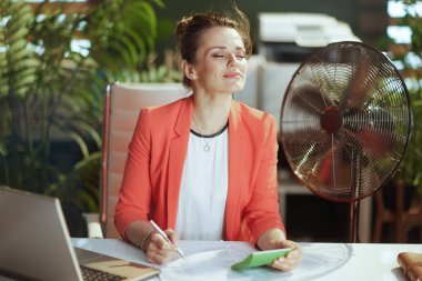 Sürdürülebilir iş yeri. Gülümseyen 40 yaşındaki modern muhasebeci kadın. Kırmızı ceketli, hesap makinesi, elektrikli vantilatör ve dizüstü bilgisayarlı..