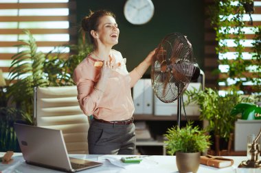 Sürdürülebilir iş yeri. Modern yeşil ofisteki 40 yıllık mutlu muhasebeci kadın. Elektronik fan ve dizüstü bilgisayarı var..