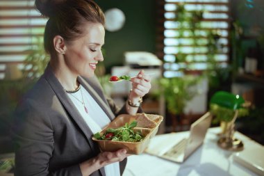 Sürdürülebilir iş yeri. Modern yeşil ofiste gri takım elbiseli, dizüstü bilgisayarı salata yiyen orta yaşlı, mutlu bir iş kadını..