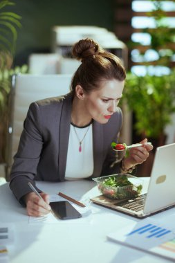 Sürdürülebilir iş yeri. Modern yeşil ofisteki gri takım elbiseli 40 yaşındaki modern iş kadını. Laptop yiyor ve akıllı telefon uygulamaları kullanıyor..