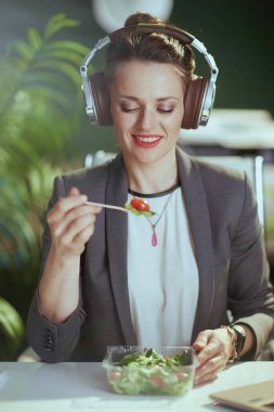 Sürdürülebilir iş yeri. Modern yeşil ofisteki gri takım elbiseli, kulaklıklı, salata yiyen mutlu modern muhasebeci kadın..