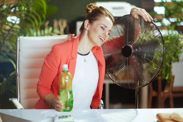 持続可能な職場 現代的なビジネス女性を笑顔で水と電気のファンのボトルと赤いジャケットで仕事中 — ストック写真