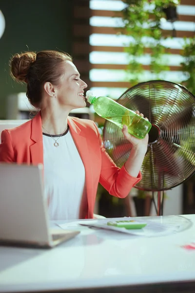 持続可能な職場 幸せな現代40歳ビジネス女性で現代緑のオフィスで赤いジャケットにボトルの水と電気ファン — ストック写真