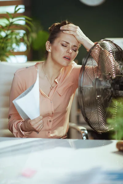 持続可能な職場 文書や夏の暑さに苦しむ電気ファンと現代の緑のオフィスで現代的な中年女性従業員 — ストック写真