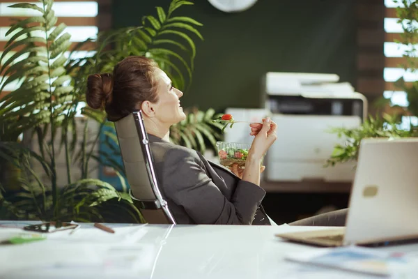 持続可能な職場 現代のグリーンオフィスのグレーのビジネススーツを着たリラックスした現代の女性従業員ラップトップでサラダを食べる — ストック写真