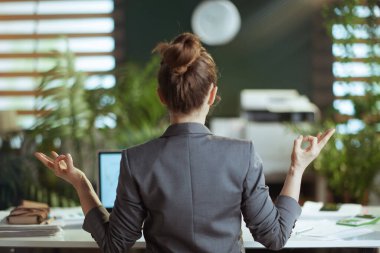Sürdürülebilir iş yeri. Modern yeşil ofiste meditasyon yapan gri takım elbiseli modern iş kadınının arkasından görüldü..