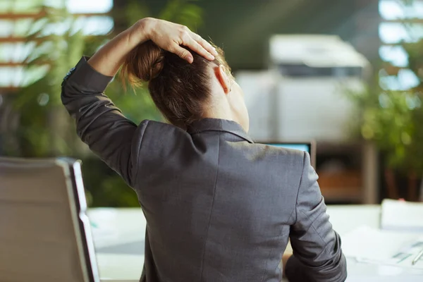 Sustainable workplace. Seen from behind modern business woman in a grey business suit at work stretching neck.