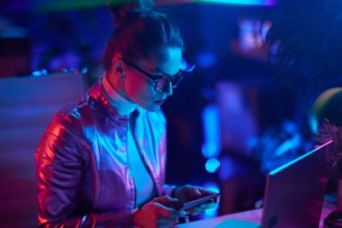 Neon metaevren fütüristik konsepti. Modern ofiste akıllı telefon kullanan gözlüklü zarif bir kadın..