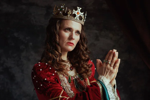 medieval queen in red dress with crown praying on dark gray background.
