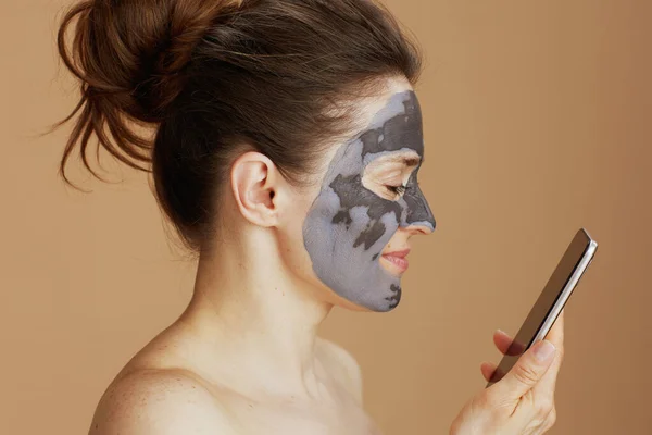 快乐的现代中年女性 戴着化妆品面具 用米色背景隔离的智能手机 — 图库照片