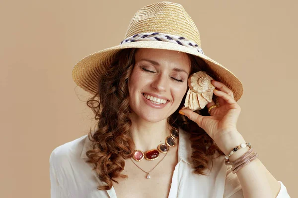 ビーチ休暇 幸せな現代主婦でホワイトブラウスとショートパンツにベージュ背景とともに麦わら帽子聞いて貝殻 — ストック写真