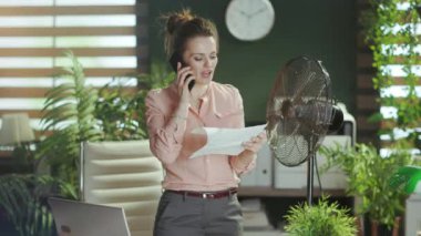 Sürdürülebilir iş yeri. Gülümseyen 40 yaşındaki muhasebeci kadın belgelerle, elektrikli vantilatörle ve bilgisayarla akıllı telefondan konuşuyor..